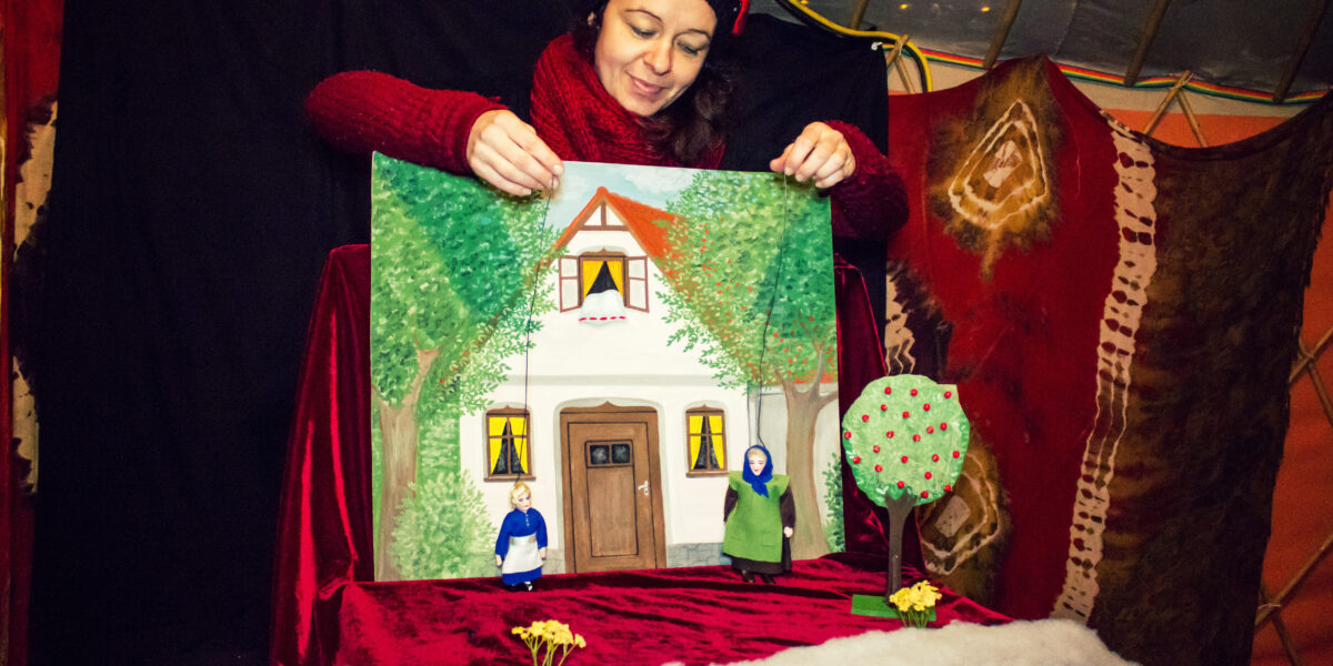 Eine Frau mit rotem Pullover und schwarzem Hut hält eine gemalte Tafel mit einem Haus, vor dem kleine Figuren und ein Modellbaum stehen, in einem zeltartigen Gebäude.