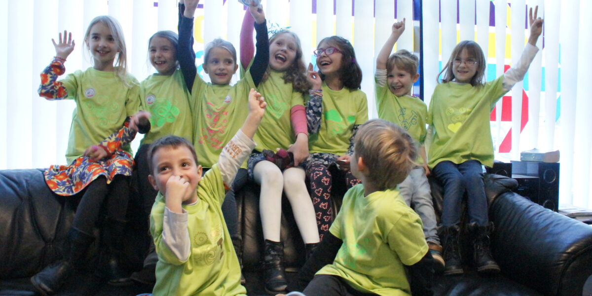 Eine Gruppe von zehn Kindern in passenden grünen T-Shirts sitzt auf einer schwarzen Couch, lächelt und hebt aufgeregt die Arme vor den vertikalen Jalousien.