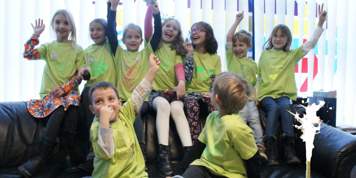 Eine Gruppe von Kindern in grünen Hemden sitzt und steht aufgeregt auf einer Couch, einige mit erhobenen Armen, vor einem Fenster. Im Vordergrund steht eine Geburtstagstorte mit einer funkelnden Kerze.