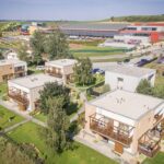 Luftaufnahme von mehreren modernen zweistöckigen Häusern mit Balkonen, grünen Rasenflächen und einem großen Gebäudekomplex mit Parkplätzen im Hintergrund.