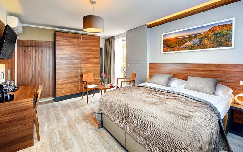 Modernes Hotelzimmer mit großem Bett, Holzmöbeln, Schreibtisch, an der Wand montiertem Fernseher, kleinem Tisch mit zwei Stühlen und einem Landschaftsgemälde über dem Bett.