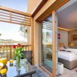 Ein Hotelzimmer mit zwei Betten öffnet sich auf einen Balkon mit einem gedeckten Tisch für das Frühstück, Orangensaft und Blumen mit Blick auf eine malerische Landschaft.