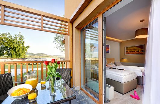 Ein Hotelzimmer mit zwei Betten öffnet sich auf einen Balkon mit einem gedeckten Tisch für das Frühstück, Orangensaft und Blumen mit Blick auf eine malerische Landschaft.