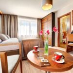 Modernes Hotelzimmer mit einem Bett, einem Schreibtisch, einem Flachbildfernseher und einem Badezimmer; auf einem runden Tisch stehen eine Vase mit rosa Blumen und zwei Tassen Kaffee. Der Raum ist von natürlichem Licht durchflutet.