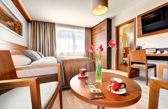 Modernes Hotelzimmer mit einem Bett, einem Schreibtisch, einem Flachbildfernseher und einem Badezimmer; auf einem runden Tisch stehen eine Vase mit rosa Blumen und zwei Tassen Kaffee. Der Raum ist von natürlichem Licht durchflutet.