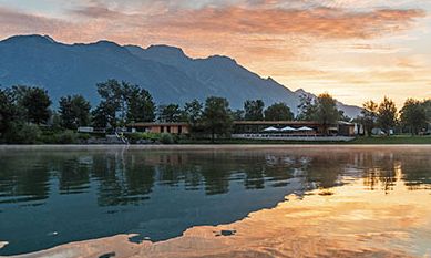 In einem ruhigen See spiegeln sich Bäume, ein Gebäude und Berge bei Sonnenuntergang, mit orangefarbenen Wolken am Himmel darüber.