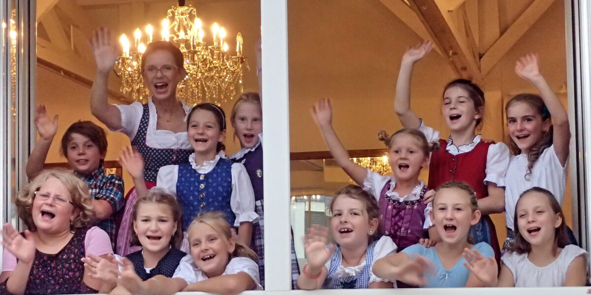 Eine Gruppe von Kindern und ein Erwachsener, alle in traditioneller Kleidung, lächeln und winken aus einem offenen Fenster, in dem Kronleuchter zu sehen sind.