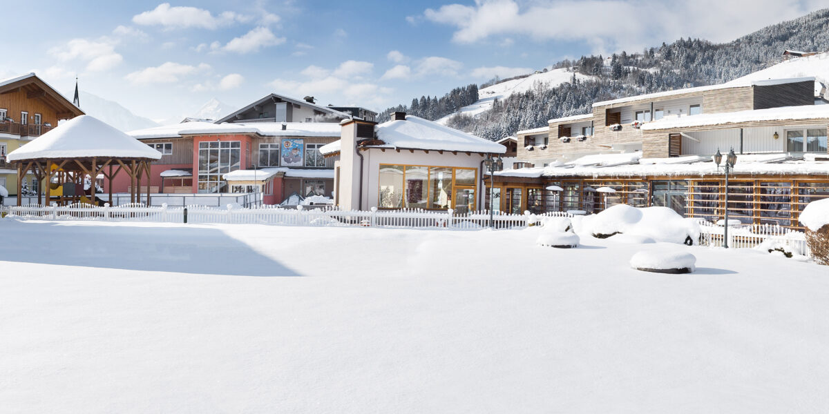 Eine schneebedeckte Freifläche vor modernen Gebäuden und Bergen unter einem teilweise bewölkten Himmel.