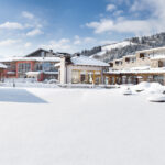 Eine schneebedeckte Freifläche vor modernen Gebäuden und Bergen unter einem teilweise bewölkten Himmel.