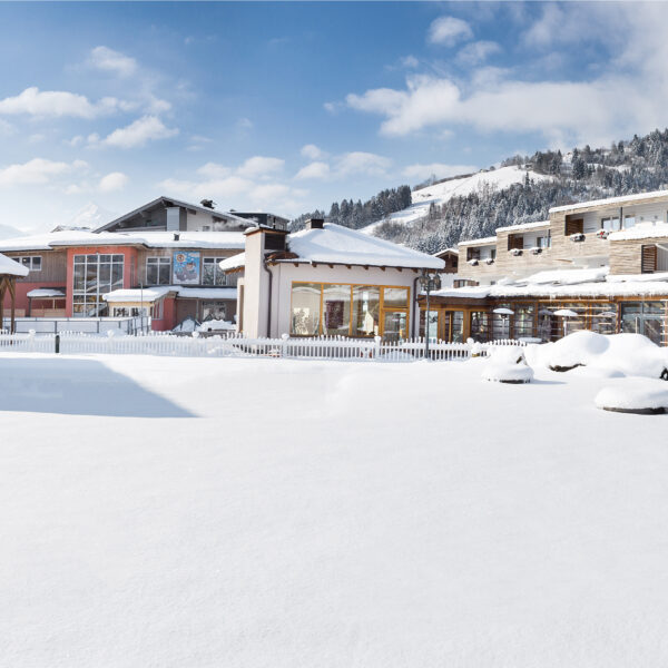 Eine schneebedeckte Freifläche vor modernen Gebäuden und Bergen unter einem teilweise bewölkten Himmel.