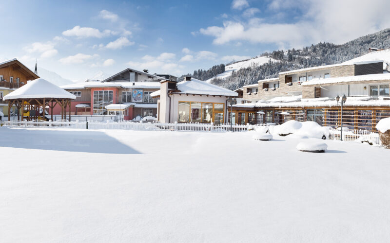 Eine schneebedeckte Freifläche vor modernen Gebäuden und Bergen unter einem teilweise bewölkten Himmel.