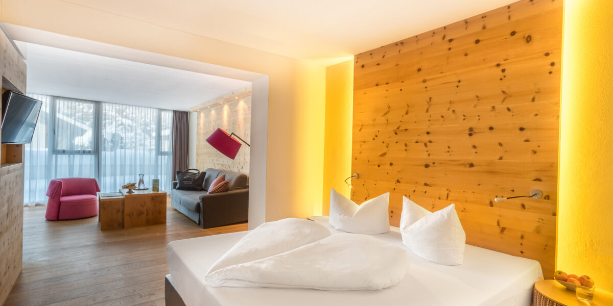 Modernes Hotelzimmer mit einem Doppelbett, einer Akzentwand aus Holz und einer separaten Sitzecke mit einem Sofa, einem rosa Sessel und einem großen Fenster mit Tageslicht.
