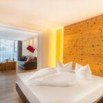 Modernes Hotelzimmer mit einem Doppelbett, einer Akzentwand aus Holz und einer separaten Sitzecke mit einem Sofa, einem rosa Sessel und einem großen Fenster mit Tageslicht.