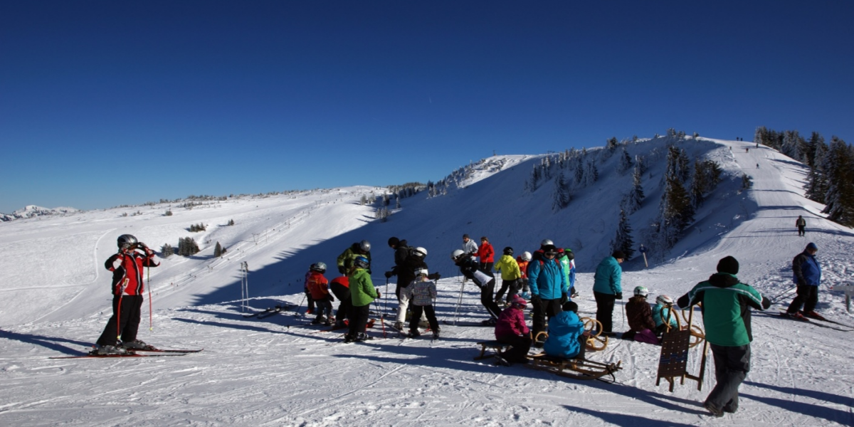 Eine Gruppe von Menschen in Winterkleidung steht mit Skiern und Schlitten an einem verschneiten Berghang unter einem klaren blauen Himmel.