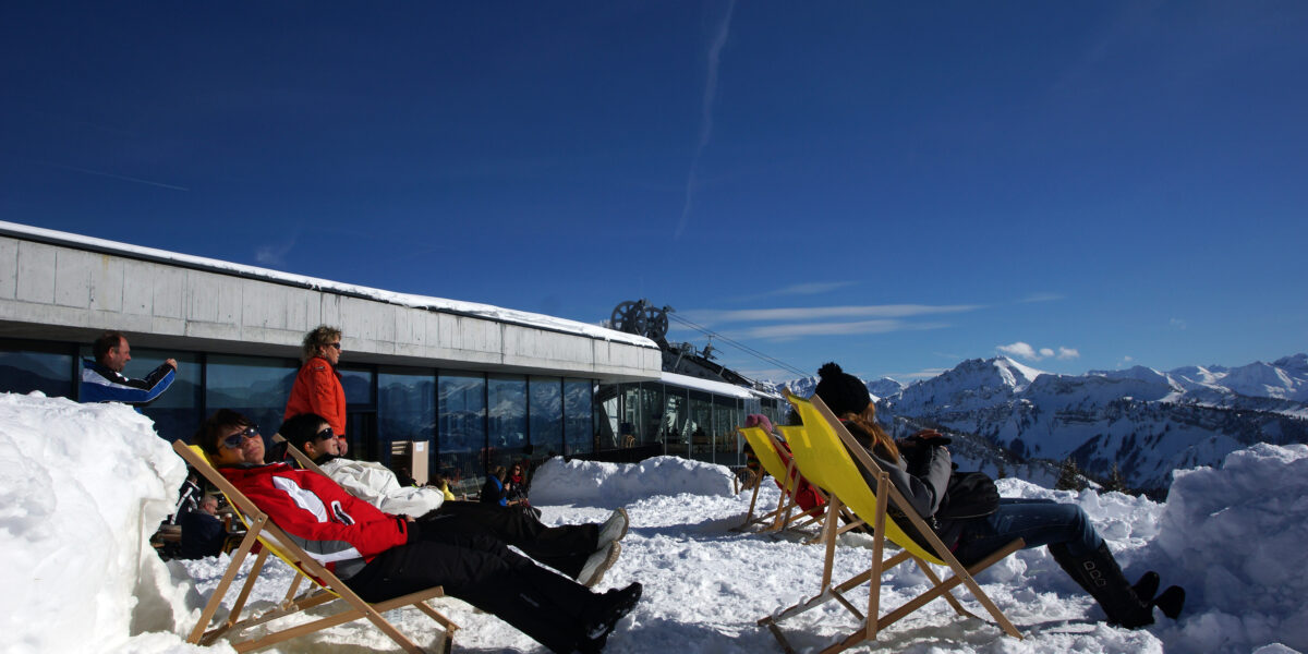 Menschen entspannen sich auf Liegestühlen im Schnee vor einem Gebäude, mit klarem Himmel und verschneiten Bergen im Hintergrund.