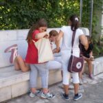 Vier Mädchen stehen und sitzen auf einer Steinbank im Freien, schauen in Taschen und unterhalten sich. Im Hintergrund sind Grünpflanzen und ein Metallzaun zu sehen.