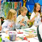 Drei kleine Kinder in mit Farbe bespritzten Kitteln konzentrieren sich auf das Malen und Zeichnen an einem Tisch in einem Kunstunterrichtsraum, umgeben von Kunstzubehör.