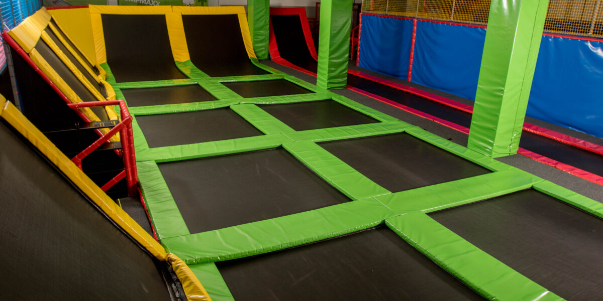 Ein Indoor-Trampolinpark mit miteinander verbundenen rechteckigen Trampolinen, die mit bunten Polstern eingefasst und von Sicherheitsnetzen umgeben sind.