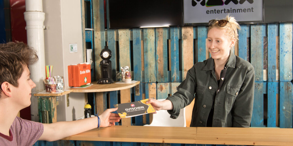 Eine Frau steht hinter einem Tresen und überreicht einem Mann an einem Stand mit der Aufschrift "MAXX entertainment" einen Gutschein, der vor einer rustikalen Holzkulisse liegt.