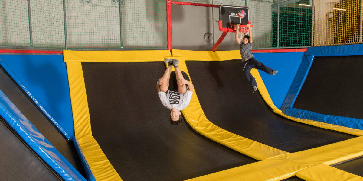 Zwei Kinder spielen auf einem Hallentrampolin; das eine ist kopfüber mitten im Sprung, das andere springt in Richtung eines Basketballkorbs.