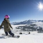 Person, die an einem sonnigen Tag einen verschneiten Berghang hinunterfährt, mit schneebedeckten Gipfeln und klarem blauen Himmel im Hintergrund.