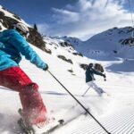 Zwei Personen fahren eine frisch präparierte Piste in einer verschneiten Berglandschaft mit Skiliften und felsigen Gipfeln im Hintergrund hinunter.