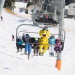 Skifahrer fahren mit einem Sessellift einen verschneiten Berg hinauf; eine Person in der Gruppe trägt ein leuchtend gelbes Tierkostüm.
