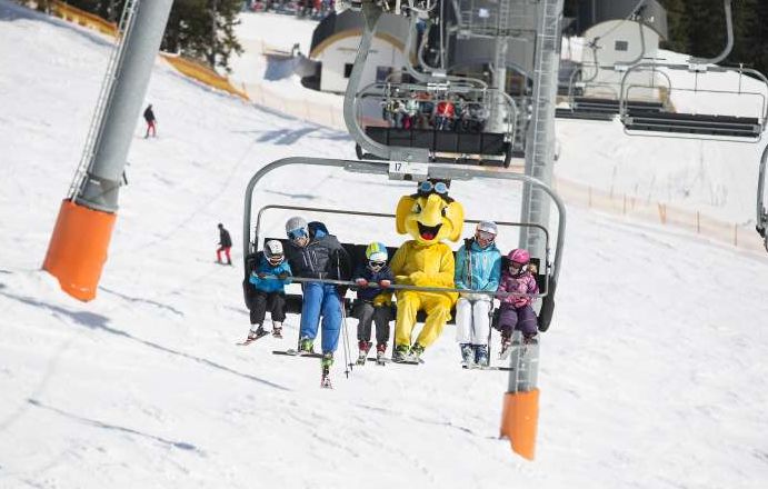 Skifahrer fahren mit einem Sessellift einen verschneiten Berg hinauf; eine Person in der Gruppe trägt ein leuchtend gelbes Tierkostüm.