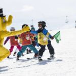 Eine Person in einem gelben Elefantenkostüm begrüßt eine Gruppe von Kindern, die auf einer verschneiten Piste das Skifahren lernen.