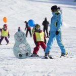 Kinder und ein Skilehrer fahren auf einer verschneiten Piste Ski. Sie tragen Helme und bunte Westen, im Hintergrund sind Cartoon-Tierausschnitte und Luftballons zu sehen.