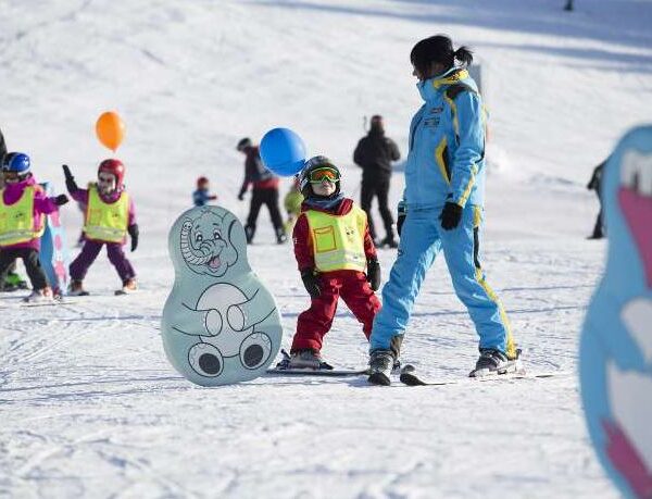 Kinder und ein Skilehrer fahren auf einer verschneiten Piste Ski. Sie tragen Helme und bunte Westen, im Hintergrund sind Cartoon-Tierausschnitte und Luftballons zu sehen.