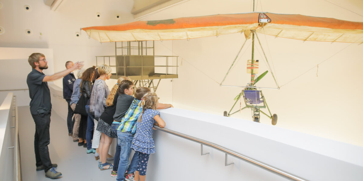 Eine Gruppe von Kindern und Erwachsenen steht an einem Geländer und betrachtet ein großes Segelflugzeug, das in einem hellen, modernen Museumsraum ausgestellt ist.