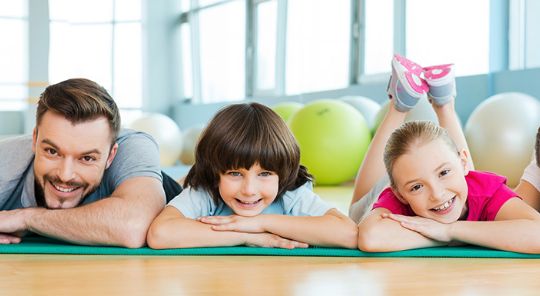 Ein Mann und zwei Kinder liegen auf Turnmatten in einem Fitnessstudio und lächeln in die Kamera, während im Hintergrund Fitnessbälle zu sehen sind.