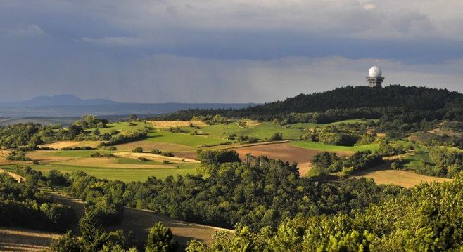 Wogende grüne und braune Felder mit Baumgruppen, ein bewaldeter Hügel und eine große weiße Radarkuppel, die auf dem Hügel unter einem bewölkten Himmel zu sehen ist.