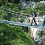 Drei Personen überqueren eine schmale Hängebrücke über eine felsige Schlucht mit Grünpflanzen und einem Wasserfall im Hintergrund.