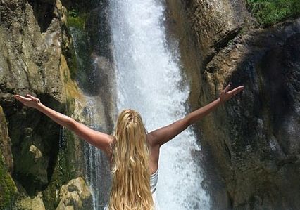 Eine Person mit langem blondem Haar steht mit ausgestreckten Armen vor einem Wasserfall, der über felsiges Gelände stürzt.