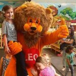 Eine Person in einem Löwenmaskottchenkostüm mit der Aufschrift "LEO" posiert mit lächelnden Kindern in einem mit Cartoon-Löwen dekorierten Indoor-Spielbereich.