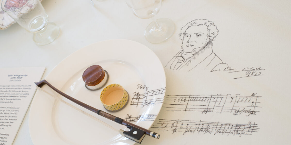 Auf einem weißen Tischtuch steht ein Teller mit zwei Desserts und einem Geigenbogen, daneben die Skizze eines Mannes, eine handschriftliche Unterschrift und Musiknoten. Im Hintergrund stehen leere Gläser.