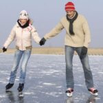 Zwei Menschen, die auf einem zugefrorenen See Schlittschuh laufen, halten sich an den Händen und tragen Winterkleidung, während im Hintergrund der Himmel klar ist.