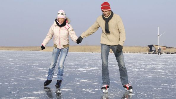 Zwei Menschen, die auf einem zugefrorenen See Schlittschuh laufen, halten sich an den Händen und tragen Winterkleidung, während im Hintergrund der Himmel klar ist.