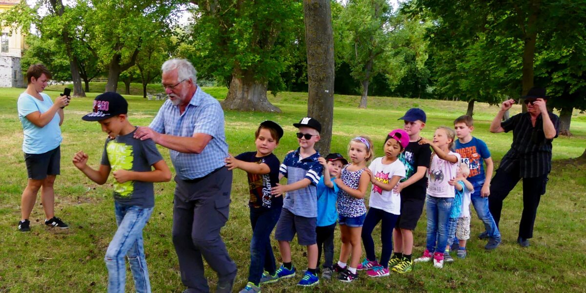Eine Gruppe von Kindern und ein älterer Mann bilden eine Conga-Linie in einem grasbewachsenen Park, während zwei Erwachsene im Hintergrund Fotos machen.