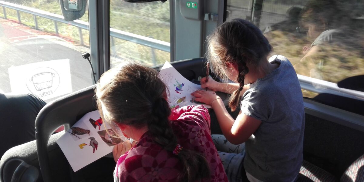 Zwei Kinder mit geflochtenen Haaren sitzen in einem Bus und zeichnen und malen Bilder auf Papier, während das Sonnenlicht durch die Fenster strömt.