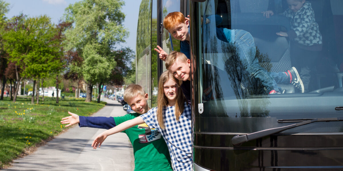 Vier Kinder lehnen sich lächelnd und gestikulierend aus einem geparkten Bus, während im Hintergrund Bäume und ein Bürgersteig zu sehen sind.