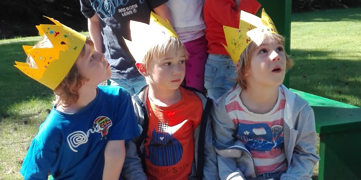 Sechs kleine Kinder, die gelbe Papierkronen tragen, sitzen und stehen auf einer grünen Bank im Freien und blicken alle nach oben zu etwas, das nicht zu sehen ist.
