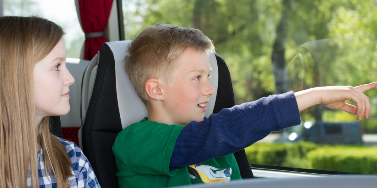 Zwei Kinder sitzen nebeneinander in einem Bus; der Junge zeigt aus dem Fenster, während das Mädchen ihn anschaut.
