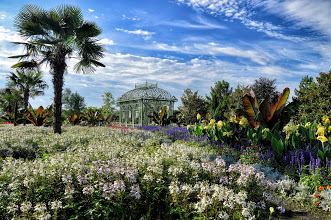 Ein Glaspavillon steht in einem Garten voller blühender Blumen, tropischer Pflanzen und einer Palme unter einem teilweise bewölkten Himmel.