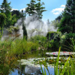 Ein üppiger, grüner Garten mit einem Teich mit Seerosenblättern, umgeben von hohen Gräsern und Bäumen, mit aufsteigendem Nebel im Hintergrund unter einem teilweise bewölkten Himmel.