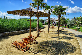 Unter strohgedeckten Sonnenschirmen und Palmen stehen hölzerne Liegestühle auf einer Sandfläche neben einem Steinweg, mit hohem Gras und blauem Himmel im Hintergrund.