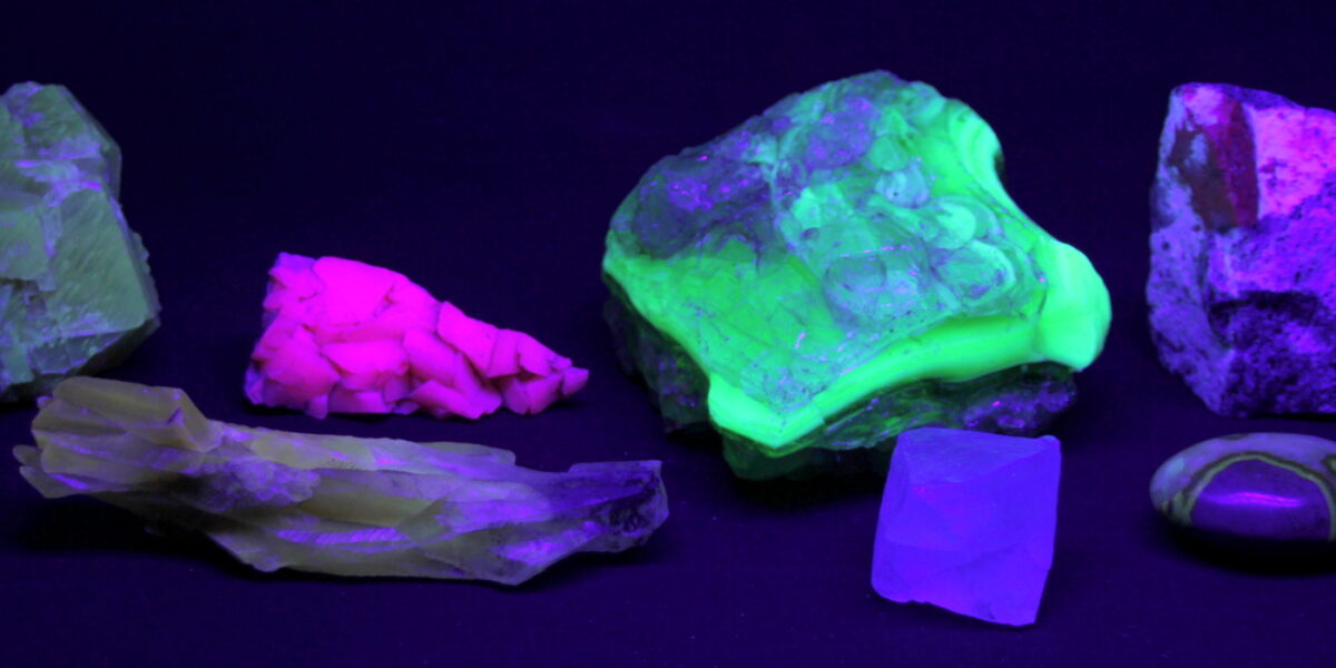 Eine Sammlung von Mineralien, die unter ultraviolettem Licht in verschiedenen Neonfarben leuchten, darunter leuchtend grüne, rosa und violette Farbtöne.