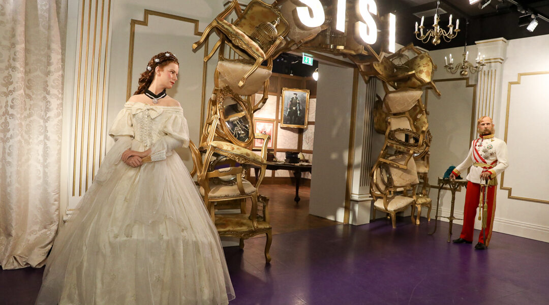 In einem Wachsfigurenkabinett sind die Figuren einer Frau in einem weißen Kleid und eines Mannes in Uniform zu sehen. Im Hintergrund befindet sich ein Bogen aus Stühlen und ein "SISI"-Schild.